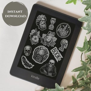 May include: A black and white digital reader with a black background and a variety of black and white stickers on the screen. The stickers include a skull, a frog, a castle, a feather, a book, a skeleton, and a globe. The reader has the word "kindle" printed on the bottom.