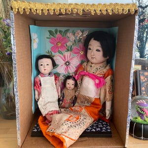 以下が含まれることがあります： 段ボール箱の中に飾られた3体の日本人形。人形は伝統的な衣装を着ており、1体はオレンジ色の着物を着ています。背景にはピンクの花のフローラルデザインがあります。箱は金色の生地でトリミングされています。