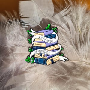 Könnte beinhalten: Ein farbenfroher Emaille-Pin mit einem Stapel Bücher mit lila und blauen Einbänden. Ein weißes Banner mit dem Text "ASK ME ABOUT MY TO BE READ PILE" umschlingt die Bücher. Grüne Blätter und Akzente vervollständigen das Design.