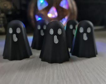 Könnte beinhalten: Eine Ansammlung kleiner, schwarzer Geisterfiguren mit weißen Gesichtern. Die Geister sind vor einer beleuchteten Kürbisdekoration angeordnet, was ein Halloween-Thema erzeugt. Der Hintergrund ist unscharf, wodurch die Aufmerksamkeit auf die Figuren gelenkt wird.