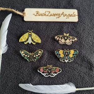 Könnte beinhalten: Fünf Emaille-Pins mit verschiedenen Mottiv-Designs. Die Motten sind schwarz, gelb, grün und pink mit goldenen Akzenten. Die Pins befinden sich auf einem schwarzen Hintergrund mit einer weißen Feder.