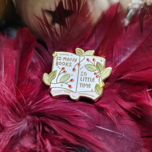 Könnte beinhalten: Ein weiß-goldener Emaille-Pin mit einem offenen Buch-Design mit dem Text "SO MANY BOOKS" und "SO LITTLE TIME". Der Pin ist mit grünen Blättern und roten Beeren verziert. Der Pin ist vor einem Hintergrund aus burgunderfarbenen Federn platziert.