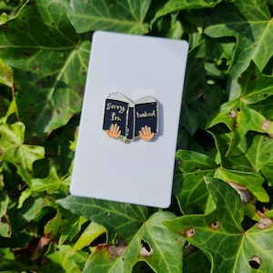 Könnte beinhalten: Schwarze Emaille-Anstecknadel in Form eines geöffneten Buches mit den Worten "Sorry I'm booked" auf den Seiten. Die Anstecknadel hat goldene Akzente und einen goldenen Verschluss.