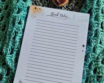 Könnte beinhalten: Ein weißes, liniertes Notizbuch mit den Worten "Book Notes" in schwarzer Kursivschrift. Das Notizbuch hat einen Aquarellhintergrund in Orange- und Blautönen und Sterne. Am unteren Rand des Notizbuchs befindet sich eine Schwarzweißillustration eines geöffneten Buches mit einer Blume.