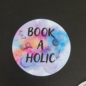 Könnte beinhalten: Ein runder Aufkleber mit einem Aquarellhintergrund in Blautönen, Lila und Rosa. Der Aufkleber zeigt den Text "BOOK A HOLIC" in schwarzer Schrift. Der Aufkleber enthält außerdem einen Halbmond, einen Schmetterling, ein Buch, eine Feder und eine Taschenuhr.
