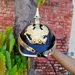 May include: A black and gold military helmet with a gold eagle emblem. The helmet has a gold spike on top and gold trim around the base. A black leather strap is attached to the helmet.