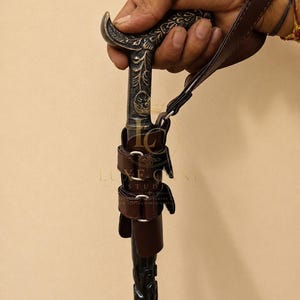 Puede incluir: Un bastón con un mango de metal oscuro adornado con intrincados grabados florales. El eje es negro con detalles tallados e incluye una correa de cuero marrón y un accesorio de cuero marrón asegurado con hebillas de metal de tono plateado.