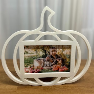 May include: White pumpkin-shaped picture frame. The frame holds a photograph of a baby surrounded by pumpkins. The frame is made of a light-colored material and sits on a wooden surface. The photo area is 4 x 6 inches (10.2 cm x 15.2 cm).