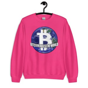 Könnte beinhalten: Ein leuchtend pinkfarbenes Sweatshirt mit einem großen Grafikdesign, das ein Bitcoin-Symbol über einem Globus zeigt. Der Text "Bitcoin Rules the World" ist in Weiß gedruckt. Das Sweatshirt hängt an einem schwarzen Kleiderbügel.