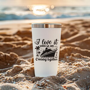 May include: White travel mug with a black design that says "I love it when we are cruising together". The design includes a cruise ship, palm trees, a sun, and an anchor. The mug is sitting on a sandy beach.