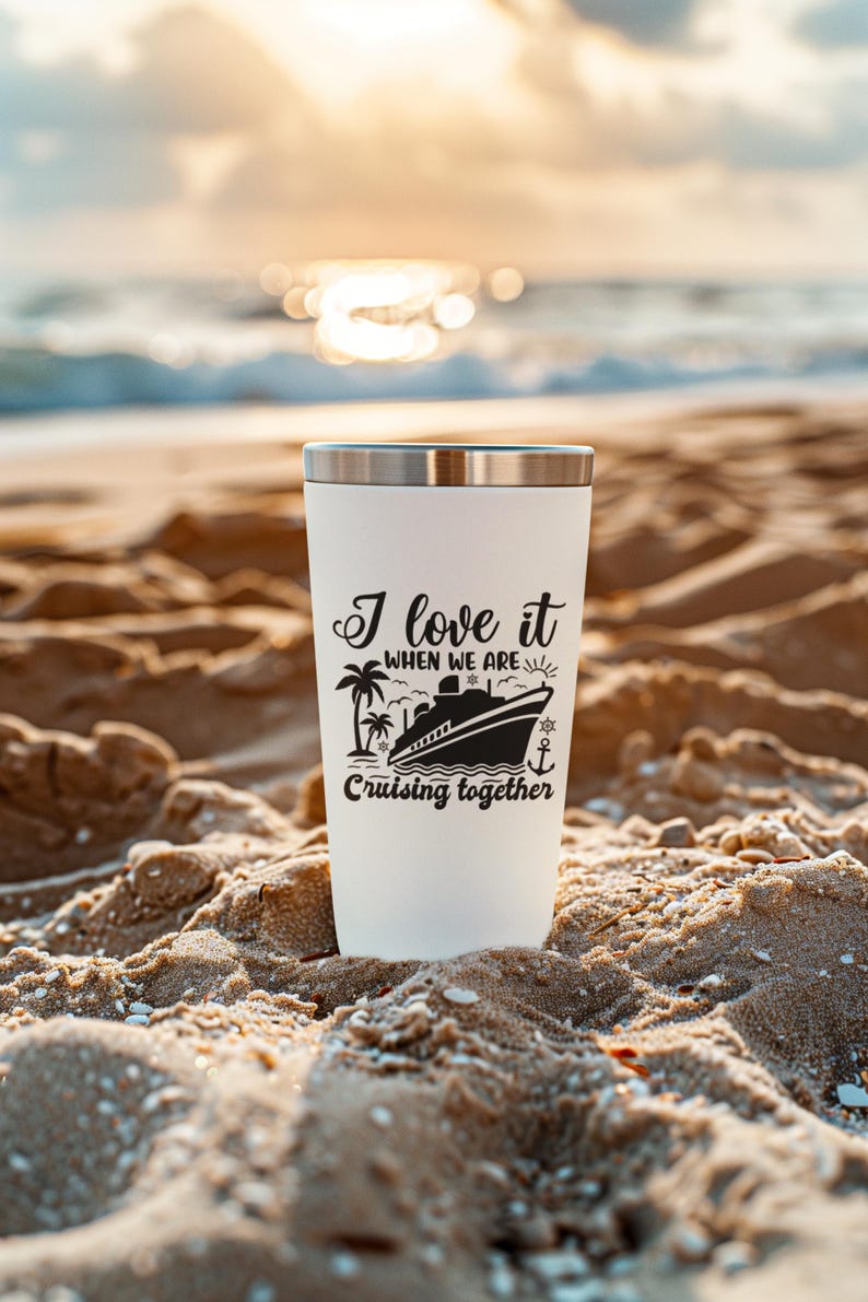 May include: White travel mug with a black design that says "I love it when we are cruising together". The design includes a cruise ship, palm trees, a sun, and an anchor. The mug is sitting on a sandy beach.