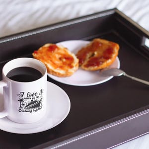 May include: White coffee mug with a black design of a cruise ship and the text "I love it when we are cruising together". The mug is on a white saucer and a tray with two halves of a bagel with jelly on top.
