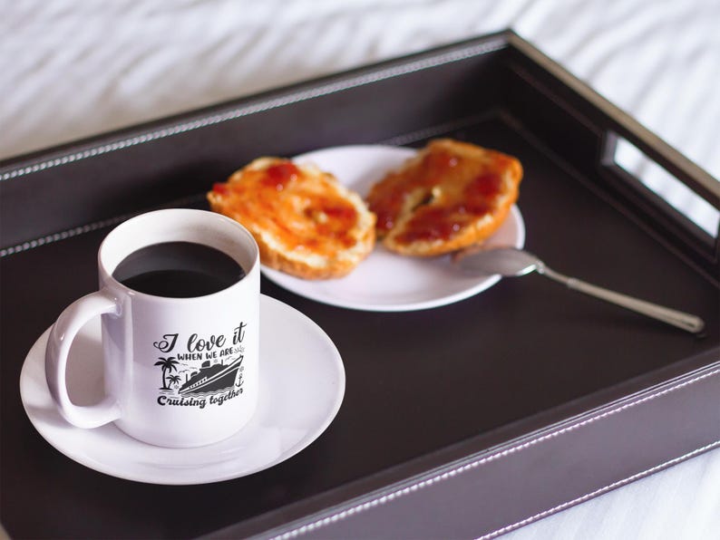 May include: White coffee mug with a black design of a cruise ship and the text "I love it when we are cruising together". The mug is on a white saucer and a tray with two halves of a bagel with jelly on top.