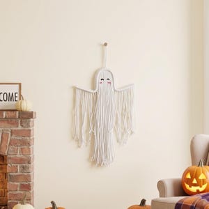 May include: A white macrame wall hanging shaped like a ghost with a rounded top, outstretched arms, and long dangling fringe. The ghost features two black embroidered eyes and two small pink embroidered cheeks on a woven cord structure.