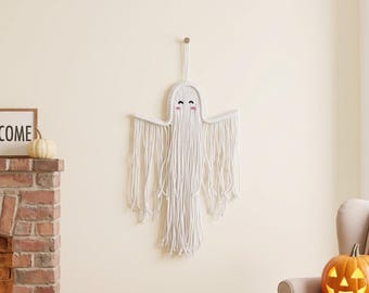 May include: A white macrame wall hanging shaped like a ghost with a rounded top, outstretched arms, and long dangling fringe. The ghost features two black embroidered eyes and two small pink embroidered cheeks on a woven cord structure.