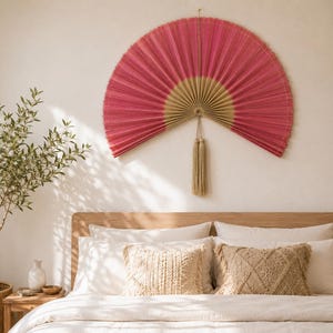May include: A large, semi-circular wall fan featuring a vibrant pink pleated fabric upper section and a natural beige base. The fan is suspended by a thin cord and includes a long, matching beige decorative tassel hanging from the center point.