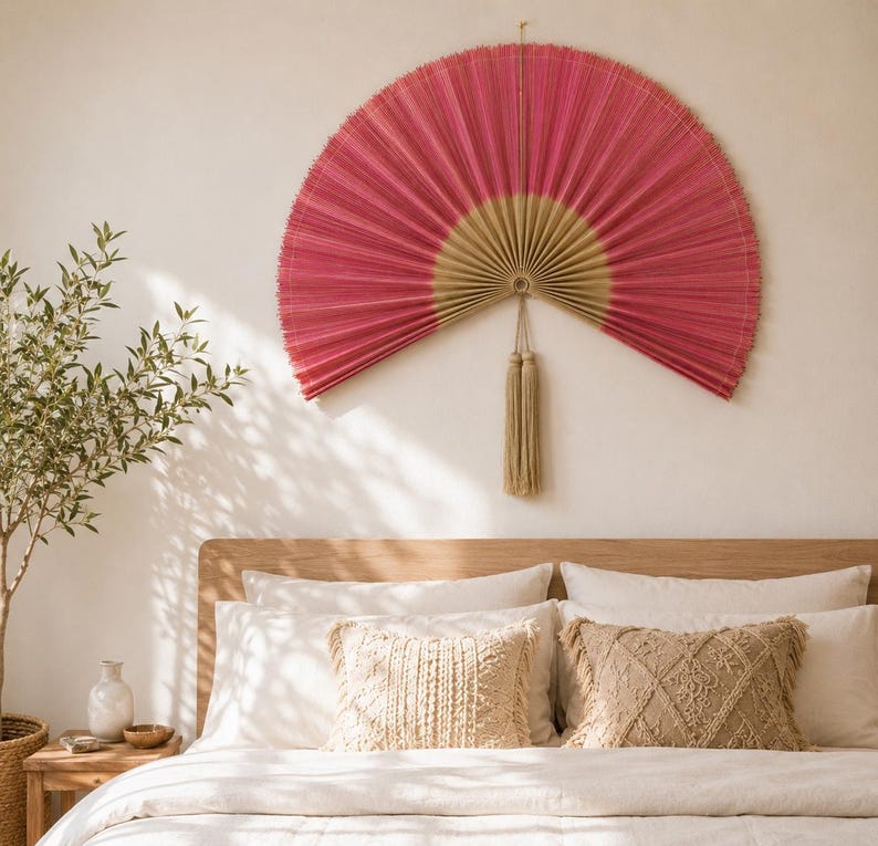 May include: A large, semi-circular wall fan featuring a vibrant pink pleated fabric upper section and a natural beige base. The fan is suspended by a thin cord and includes a long, matching beige decorative tassel hanging from the center point.