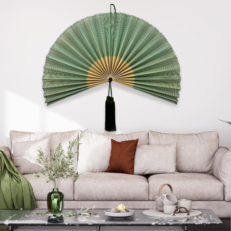 May include: A large, semi-circular wall fan featuring pleated green and gold paper panels. The fan is suspended by a thin cord and includes a decorative black tassel hanging from the center pivot point, creating a symmetrical display piece.