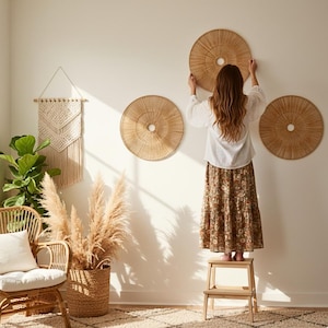 Pode incluir: Três peças redondas de decoração de parede tecidas com um orifício central são exibidas em uma parede branca. Uma pessoa está em pé em um pequeno banco de madeira, pendurando uma das peças. O quarto também apresenta uma tapeçaria de macramê, uma cadeira de vime e uma cesta de capim-dos-pampas.