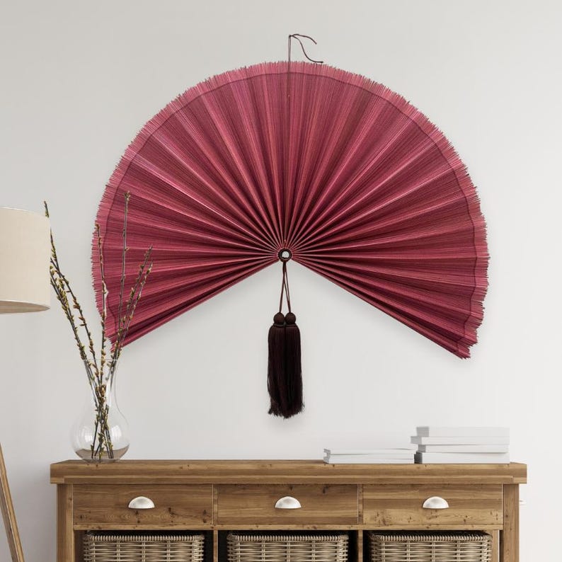 May include: A large, semi-circular wall fan in a deep burgundy red color with fine, vertical pleats. The fan features a dark brown decorative tassel hanging from the center point, suspended by a thin cord against a plain, light-colored wall.