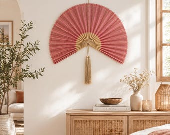 May include: A large, semi-circular wall fan featuring a pleated, deep rose-pink fabric surface with a light tan wooden base. The fan is suspended from a small loop and includes a long, matching tan decorative tassel hanging from the center point.