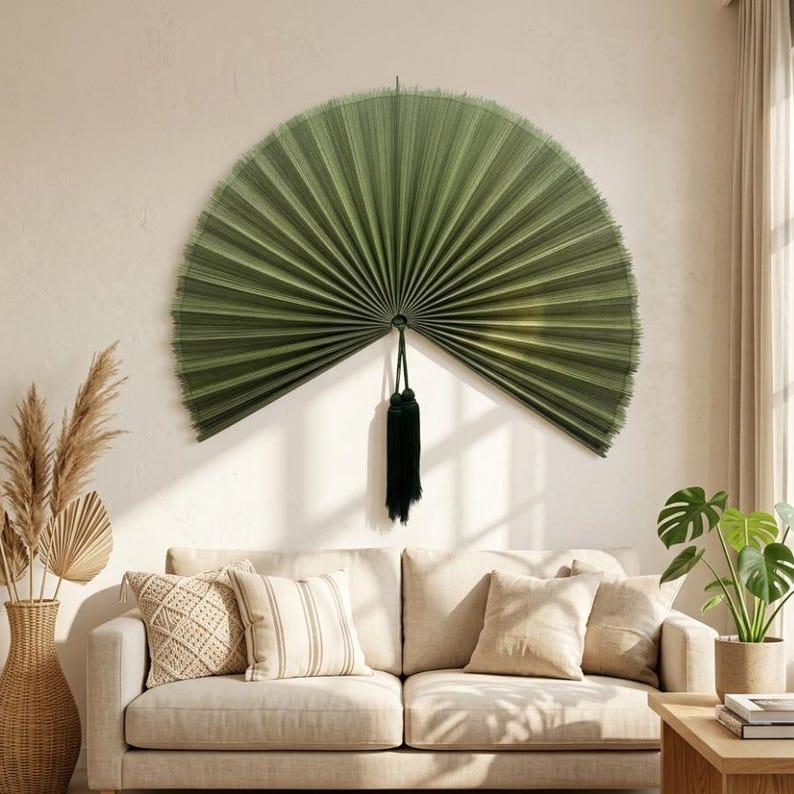May include: A large, semi-circular wall fan in a deep olive green color, featuring fine, radiating pleats and a textured, fibrous appearance. A dark green decorative tassel hangs from the center point of the fan, adding a vertical accent.