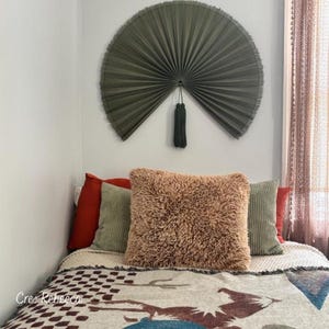 May include: A large, semi-circular olive green pleated wall fan with a central dark tassel hangs above a bed. The bed features a fluffy tan square pillow, two smaller sage green striped pillows, two red pillows, and a patterned throw blanket.