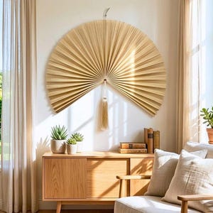 May include: A large, semi-circular wall hanging made of natural, light-colored dried palm leaves arranged in a pleated, fan-like pattern. A single, long, matching beige tassel hangs from the center point of the fan, suspended by a thin cord.