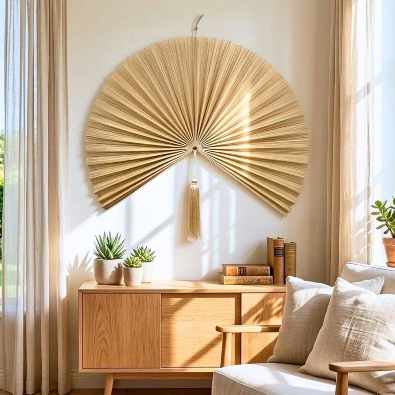May include: A large, semi-circular wall hanging made of natural, light-colored dried palm leaves arranged in a pleated, fan-like pattern. A single, long, matching beige tassel hangs from the center point of the fan, suspended by a thin cord.