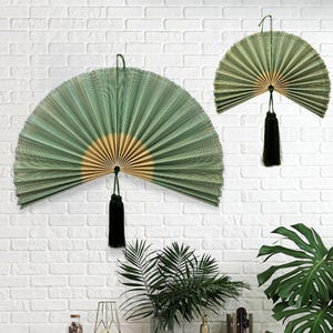 May include: Two semi-circular folding fans made of green bamboo slats with a natural wood-colored center. Each fan features a long black tassel hanging from the bottom center point, suspended against a white brick wall background.