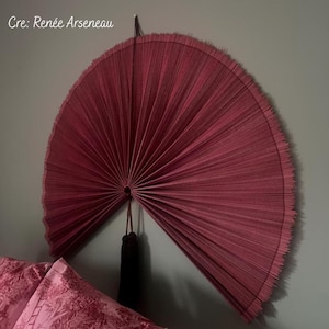 May include: A large, semi-circular wall fan in a deep burgundy red hue, featuring a pleated texture and a fringed outer edge. The fan is suspended by a thin cord and includes a dark brown tassel hanging from the central pivot point.