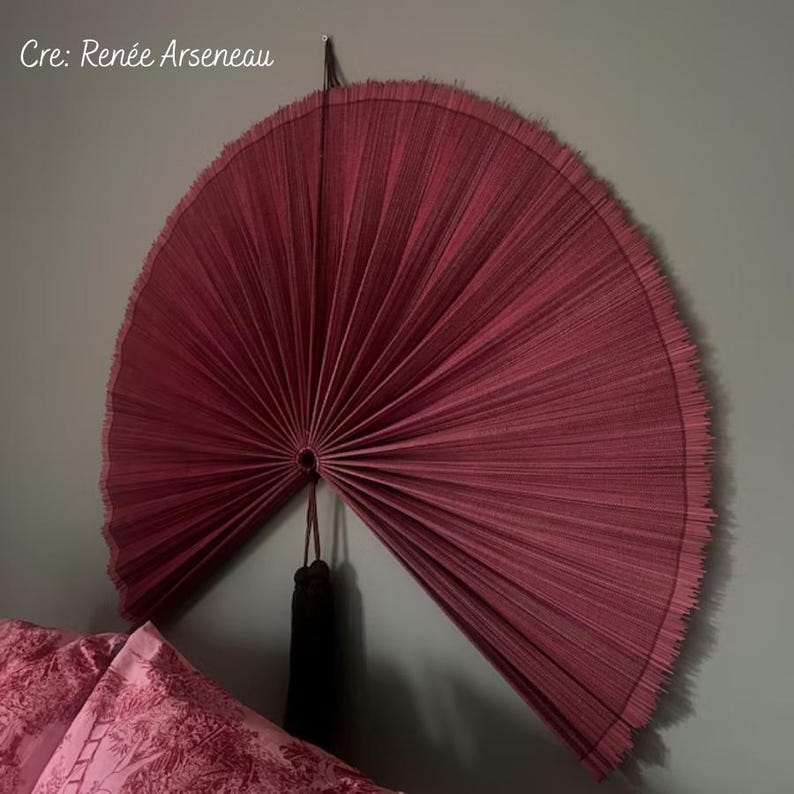 May include: A large, semi-circular wall fan in a deep burgundy red hue, featuring a pleated texture and a fringed outer edge. The fan is suspended by a thin cord and includes a dark brown tassel hanging from the central pivot point.