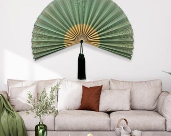 May include: A large, semi-circular wall fan featuring pleated green paper with a gold-colored center. The fan is suspended by a thin cord and features a long, black decorative tassel hanging from the central pivot point at the bottom.