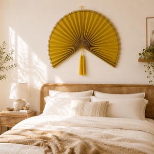 May include: A large, semi-circular mustard yellow pleated paper fan wall hanging featuring a decorative tassel suspended from the center. The fan is mounted on a light-colored wall above a bed with white bedding and a woven cane headboard.