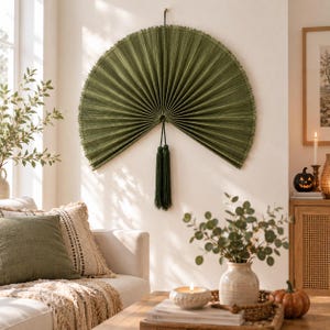 May include: A large, semi-circular wall fan in a deep olive green color, featuring a pleated texture and a frayed edge. Two long, matching green tassels hang from the center point of the fan, which is mounted securely against a plain wall.