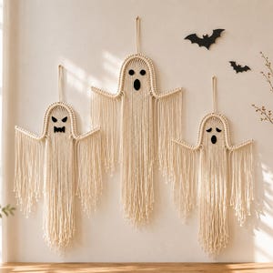 May include: Three off-white macrame ghost wall hangings with knotted cord details and long fringe. Each ghost features black felt eyes and mouths with distinct expressions. Two black paper bat cutouts are positioned near the top right corner.