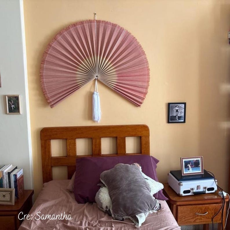May include: A large, semi-circular wall hanging fan in a dusty rose color with a central white tassel. The fan features a pleated design that radiates from a central point, creating a decorative focal point on the wall above the wooden bed frame.