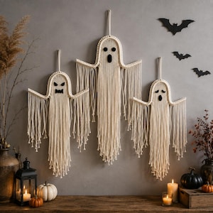 May include: Three off-white macrame ghost wall hangings with black felt eyes and mouths. Each ghost features a rounded head and long, dangling fringe cords. The ghosts are suspended by loops and display distinct facial expressions including angry, surprised, and sad.