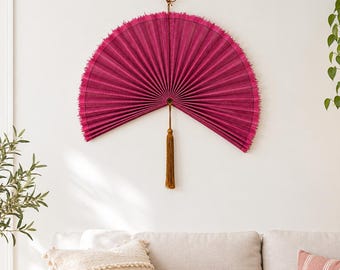 May include: A large, semi-circular wall fan in a vibrant magenta hue, featuring a pleated texture with frayed edges. A long, golden-brown decorative tassel hangs from the center point, suspended by a thin cord attached to the top.