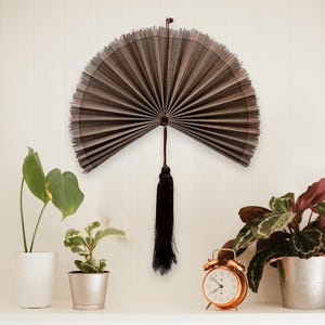 May include: A semi-circular wall fan made of thin, dark brown, vertically ribbed material. The fan features a central pivot point with a long, dark brown decorative tassel hanging downward from the base, creating a symmetrical display.