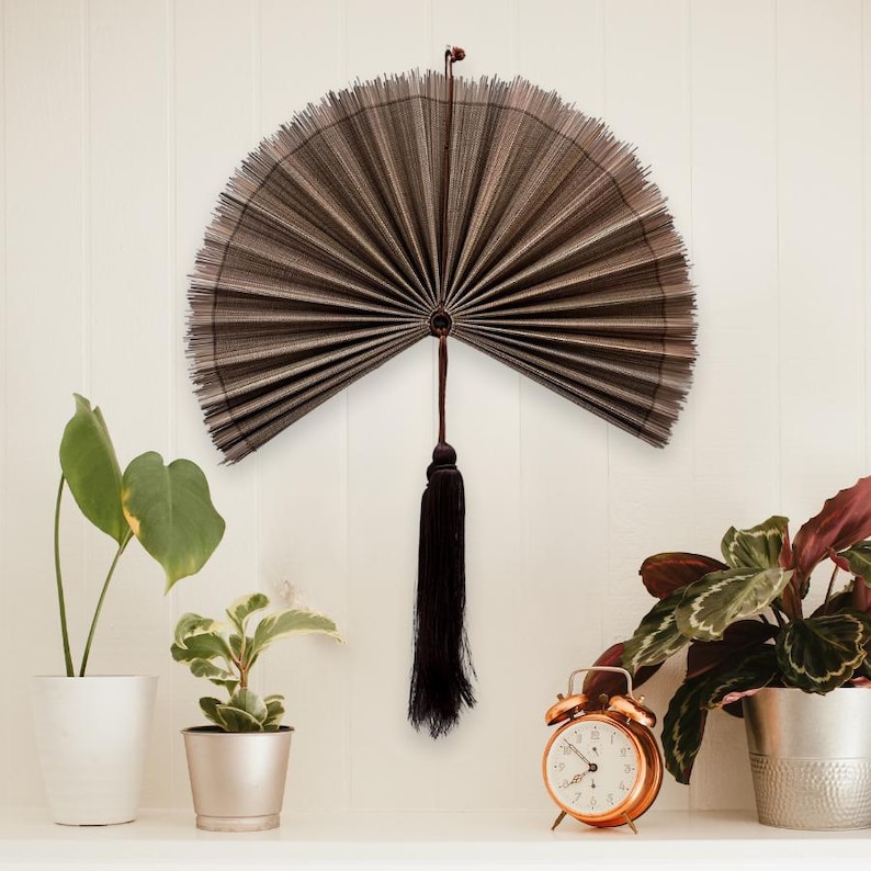 May include: A semi-circular wall fan made of thin, dark brown, vertically ribbed material. The fan features a central pivot point with a long, dark brown decorative tassel hanging downward from the base, creating a symmetrical display.