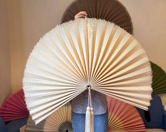 May include: A large, semi-circular hand fan made of cream-colored, finely pleated material. The fan features a central metal ring at the base with a long, matching cream-colored decorative tassel hanging from it, creating a symmetrical display.