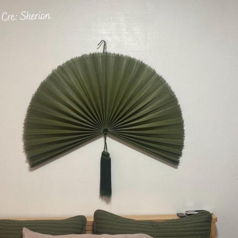 May include: A large, semi-circular wall fan made of dark olive green palm leaves, featuring a pleated texture and a matching dark green tassel hanging from the center point. The text Cre: Sherion is visible in the top left corner of the image.