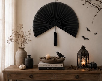 May include: A large, semi-circular black pleated paper fan wall decoration featuring a central black tassel. The fan is mounted on a wall above a wooden console table, serving as a focal point for a display of seasonal decor items.