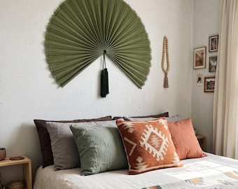 May include: A large, semi-circular olive green wall fan made of pleated palm leaves, featuring a dark green tassel hanging from the center. The fan is mounted on a white wall above a bed with various pillows and a patterned blanket.