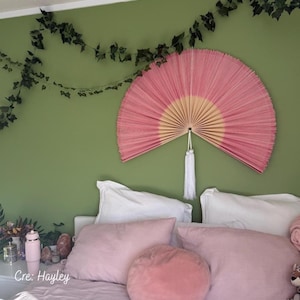 Op de afbeelding: Een grote, roze en beige waaier hangt aan een groene muur boven een bed. De waaier heeft een witte kwast en is omgeven door decoratief klimop. Het bed heeft roze en witte kussens.