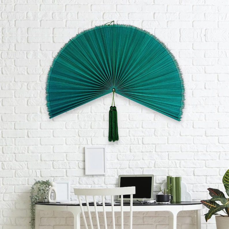 May include: A large, semi-circular wall fan in a deep teal green color. The fan features a finely pleated texture and is suspended from a central point. A matching teal green decorative tassel hangs vertically from the center of the fan base.