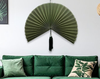 May include: A large, semi-circular wall fan in a deep olive green color, featuring a pleated texture and a fringed edge. A black decorative tassel hangs from the center point of the fan, which is mounted flat against a plain white wall.