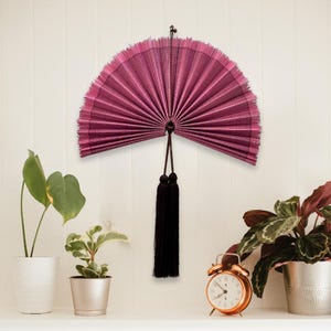 May include: A semi-circular wall-mounted folding fan in a deep magenta hue with a fine ribbed texture. The fan features a dark brown cord loop at the top for hanging and two long, dark brown decorative tassels suspended from the center base.