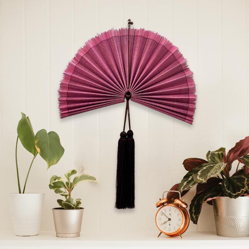 May include: A semi-circular wall-mounted folding fan in a deep magenta hue with a fine ribbed texture. The fan features a dark brown cord loop at the top for hanging and two long, dark brown decorative tassels suspended from the center base.